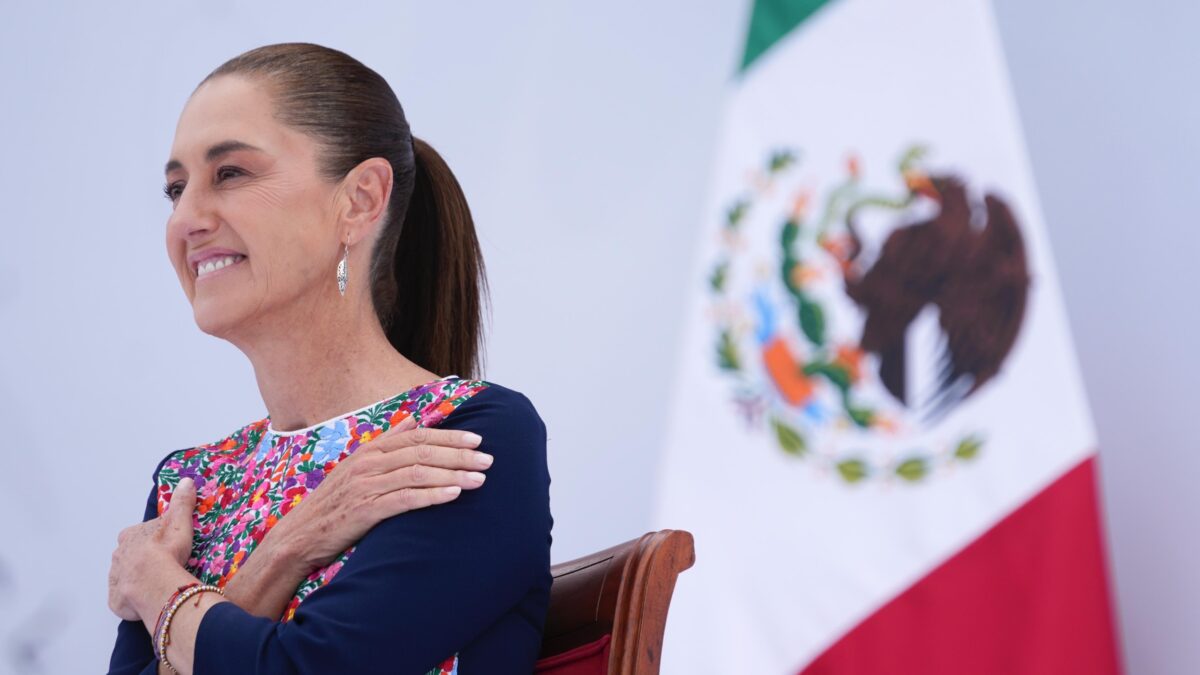 Claudia Sheinbaum sonríe sentada junto a la bandera de México durante el acto.